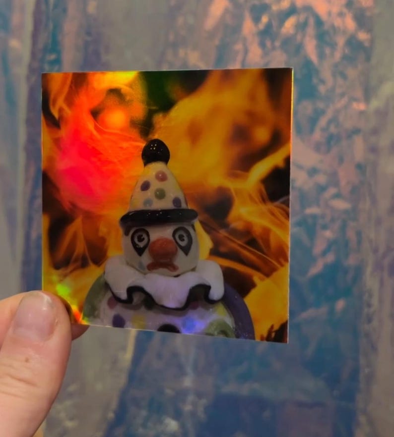 May include: A square sticker featuring a sad clown figurine with a polka-dot hat and ruffled collar. The background has a fiery, orange and yellow gradient. The sticker is held by a hand.
