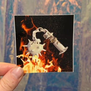 May include: A sticker featuring a white, spiky mace and a decorative, ornate tube, connected by a chain. The background shows flames against a black backdrop. The sticker is held by a person's hand.