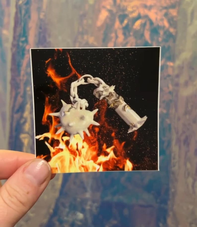 May include: A sticker featuring a white, spiky mace and a decorative, ornate tube, connected by a chain. The background shows flames against a black backdrop. The sticker is held by a person's hand.
