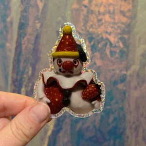 May include: A whimsical sticker featuring a clown with a red nose, hat, and strawberry-shaped body. The clown has a white face and a red and white ruffled collar. The sticker has a glittery, iridescent border.