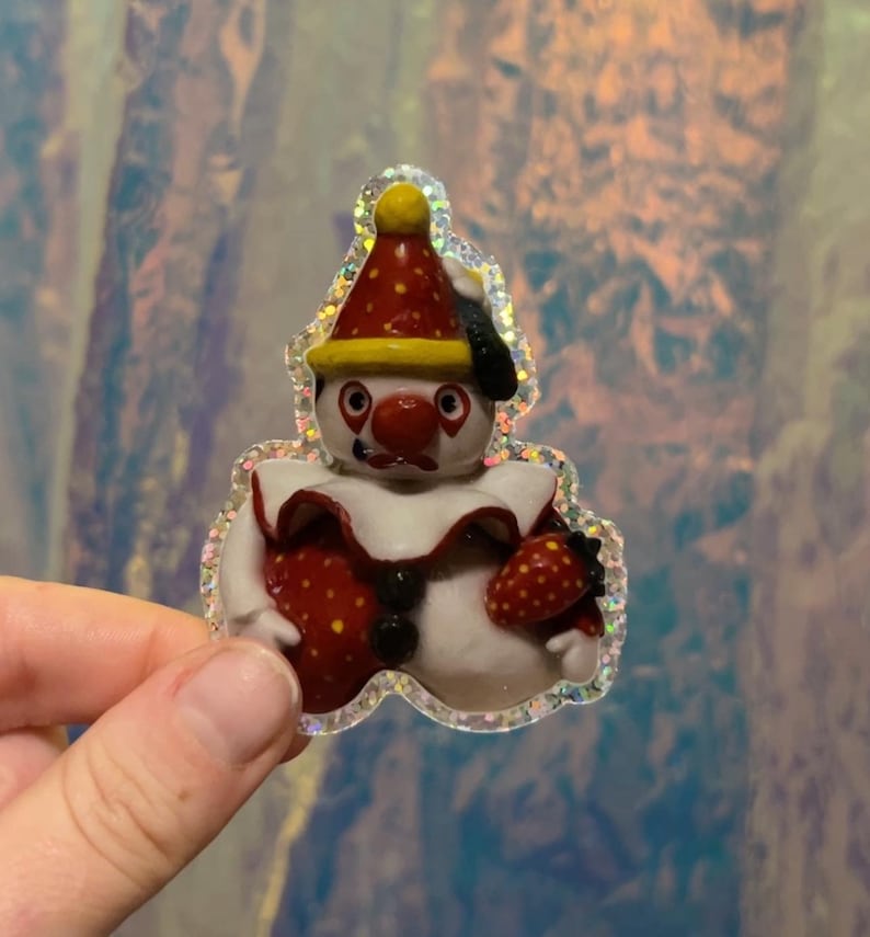 May include: A whimsical sticker featuring a clown with a red nose, hat, and strawberry-shaped body. The clown has a white face and a red and white ruffled collar. The sticker has a glittery, iridescent border.
