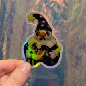 May include: A sticker featuring a sad clown with a star-topped hat, holding a crescent moon. The clown has a white face, a red nose, and a black and white collar. The sticker has a holographic effect.