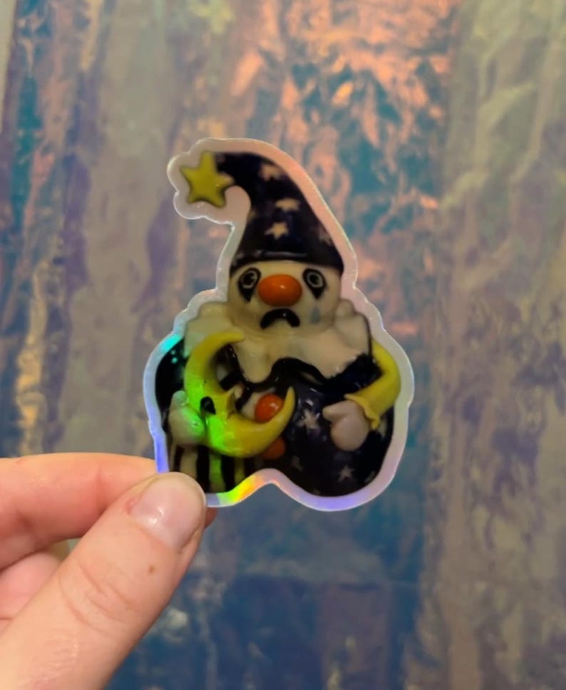May include: A sticker featuring a sad clown with a star-topped hat, holding a crescent moon. The clown has a white face, a red nose, and a black and white collar. The sticker has a holographic effect.