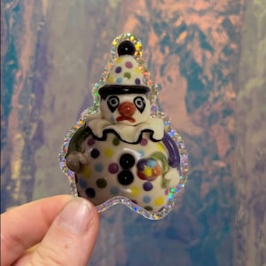 May include: A whimsical clown figurine with a sad expression. The ceramic figure features a white base with colorful polka dots, a black hat, and a ruffled collar. The figurine is outlined with a glittery border.