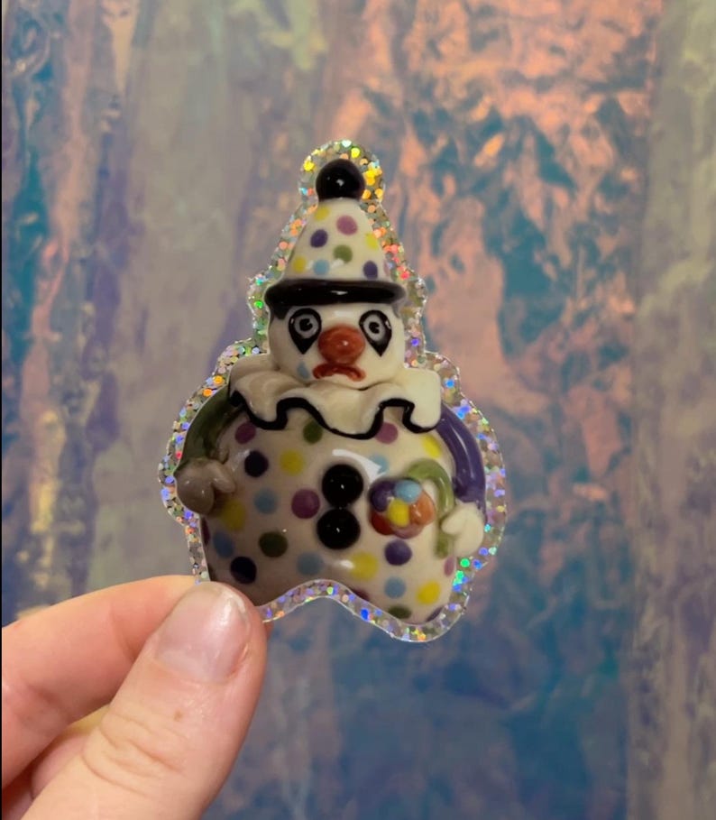 May include: A whimsical clown figurine with a sad expression. The ceramic figure features a white base with colorful polka dots, a black hat, and a ruffled collar. The figurine is outlined with a glittery border.