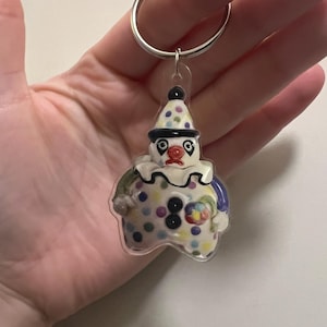 May include: A clown-shaped keychain with a silver keyring. The clown has a white base with colorful polka dots, a black hat with polka dots, and a red nose. The clown's outfit includes blue, green, and yellow accents.