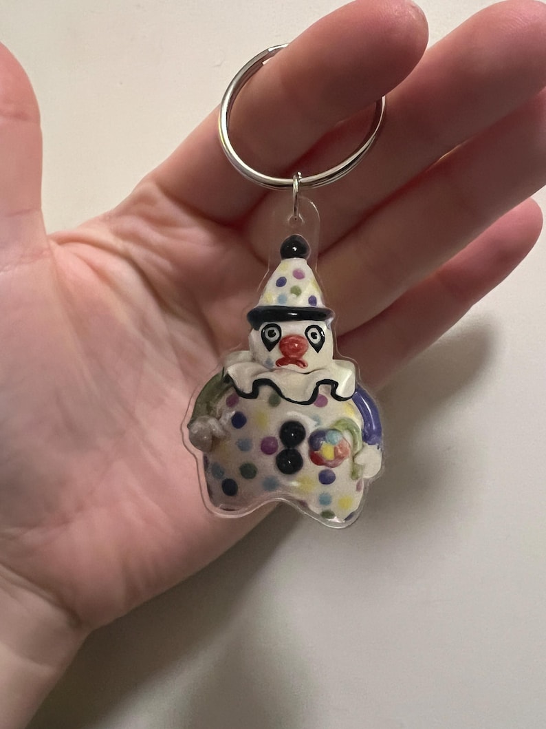May include: A clown-shaped keychain with a silver keyring. The clown has a white base with colorful polka dots, a black hat with polka dots, and a red nose. The clown's outfit includes blue, green, and yellow accents.