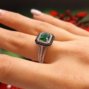 Puede incluir: Un anillo de plata con una piedra preciosa cuadrada verde esmeralda rodeada de diamantes más pequeños. El anillo tiene una banda de esmalte negro con un borde engastado con diamantes.