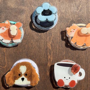 May include: A collection of five acrylic phone grips featuring cute illustrations. Designs include an orange cat, a black cat in a blue flower, another orange cat, a Cavalier King Charles Spaniel, and a smiling coffee cup.