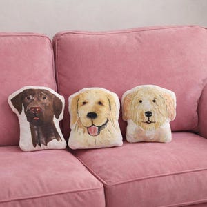 May include: Three dog-shaped throw pillows on a pink sofa. The pillows feature watercolor-style illustrations of a chocolate Labrador, a Golden Retriever, and a fluffy dog. The sofa has a soft, textured appearance.
