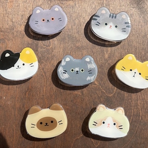 May include: Seven cat-shaped decorative magnets in various colors, including gray, yellow, black, and tan. Each magnet features a different cat face design with whiskers and eyes. The magnets are on a wooden surface.