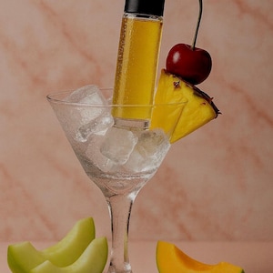May include: A cocktail-themed still life featuring a clear martini glass filled with ice cubes. A yellow liquid-filled bottle with a black cap sits in the ice. Garnishes include a cherry, pineapple slice, and melon slices. The background is a pinkish-beige.