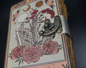 Puede incluir: Un pequeño diario encuadernado en cuero con una cubierta rosa y blanca. La cubierta presenta una ilustración de carta de tarot de Los Amantes, que representa a dos esqueletos tomados de la mano y rodeados de flores. El diario tiene un cierre de metal y una correa de cuero.