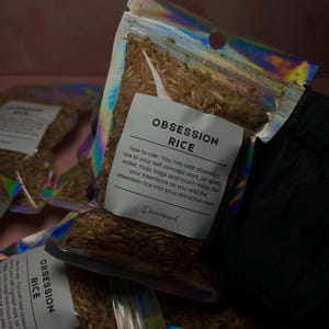 May include: Several clear, iridescent pouches filled with brown rice. Each pouch has a white label with the words "OBSESSION RICE" printed in black. Instructions for use are also printed on the label. The pouches are held by a gloved hand.