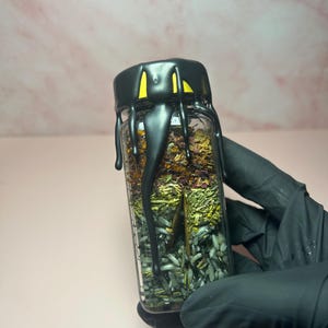 May include: A small, clear glass jar filled with layers of dried herbs and crystals. The jar has a black lid with a dripping design. The background is a soft pink.