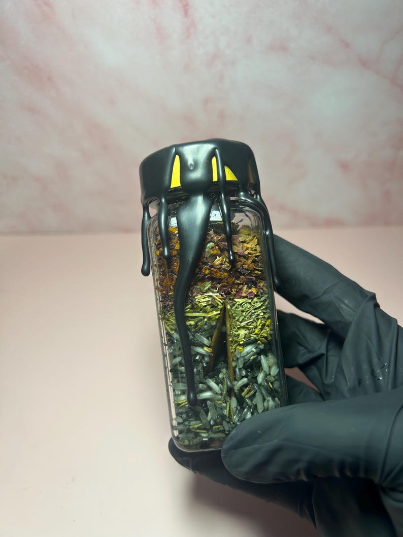 May include: A small, clear glass jar filled with layers of dried herbs and crystals. The jar has a black lid with a dripping design. The background is a soft pink.