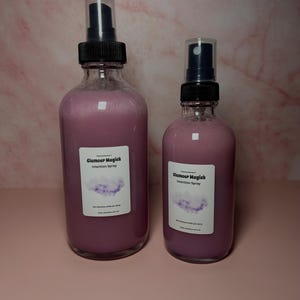 May include: Two glass spray bottles filled with a pink liquid. The bottles have black spray nozzles and white labels that read "Glamour Magick Intention Spray" with a cloud graphic. The bottles are on a pink surface.