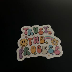 Może przedstawiać: Kolorowa naklejka z napisem "TRUST THE PROCESS" w literach bąbelkowych. Naklejka przedstawia uśmiechniętą buzię, kwiaty, tęczę i gwiazdki. Tło jest czarne.