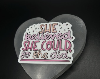 Puede incluir: Un objeto gris en forma de corazón con una pegatina blanca. La pegatina tiene el texto "SHE believed SHE COULD so SHE did" en rosa y blanco. También hay estrellas decorativas.