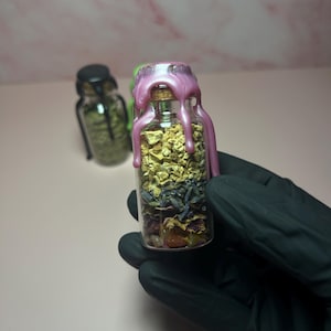 May include: A small glass bottle filled with dried herbs and crystals, sealed with a cork and pink wax. The bottle is held by a gloved hand. Another bottle with a black lid is in the background.