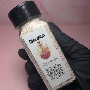 May include: A clear glass bottle with a black cap filled with beige powder, labeled "Obsession Intention Powder." The label features a heart design and text. The bottle is held by a gloved hand.