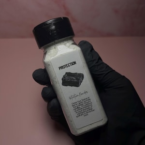 May include: A clear plastic bottle with a black lid, filled with white powder. The label reads "PROTECTION" above a graphic of a black stone and "Intention Powder". Instructions are printed below the graphic. The bottle is held by a gloved hand.