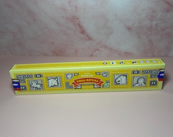 May include: A rectangular yellow box of incense sticks with the words "YOGA RITUAL" in red and gold. The box features illustrations of animals and figures. The background is a light pink marble pattern.