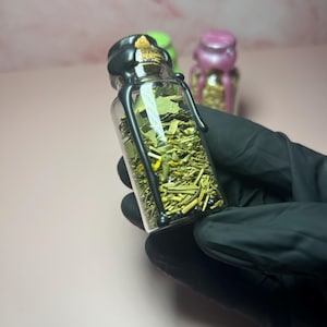 May include: A clear glass bottle filled with dried herbs and spices, sealed with a black wax top. The bottle is held by a gloved hand, with two other bottles in the background, one green and one pink.