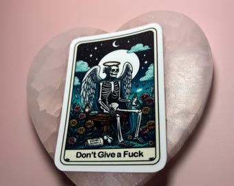 May include: A rectangular sticker featuring a skeleton with angel wings, a halo, and a moonlit night sky background. The sticker has the text "Don't Give a Fuck" at the bottom. The sticker is placed on a pink heart-shaped stone.
