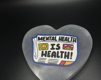 Pode incluir: Um autocolante com o texto "MENTAL HEALTH IS HEALTH!" num design em estilo jornal, com um rosto sorridente e um sapo. O autocolante é colocado numa pedra cinzenta em forma de coração.