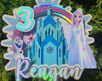 May include: A cake topper featuring the character Elsa from the movie Frozen, a blue castle, and the number 3. The name "Reagan" is also displayed. The design includes glitter and a rainbow accent.