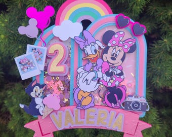 May include: A pink and blue cake topper with a rainbow, clouds, and two Disney characters, Minnie Mouse and Daisy Duck. The topper also includes a number 2, the name Valeria, and a camera.