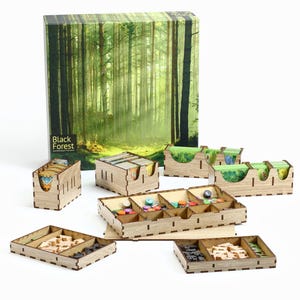 May include: Wooden organiser set for the board game Black Forest. The set includes a box, trays, and dividers for storing game components. The box features a forest scene with green trees and a path.