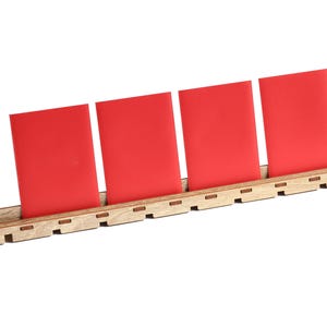 Puede incluir: Un organizador de escritorio de madera con cuatro tarjetas rojas. Las tarjetas están en posición vertical e inclinadas, sujetas por la base de madera. El organizador es de madera clara y está sobre un fondo blanco.