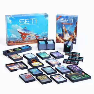Pode incluir: O conjunto de jogo de tabuleiro SETI inclui duas caixas retangulares grandes com ilustrações de temática espacial, vários organizadores de cartas em plástico preto e diversos componentes de jogo como baralhos, fichas pequenas e marcadores.
