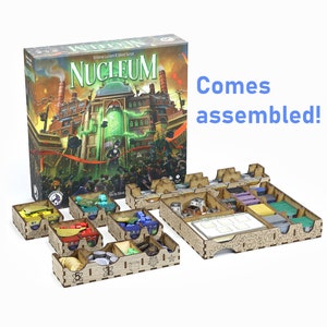 Peut inclure: Un organisateur de jeu en bois pour le jeu de société "Nucleum: Science, Lucrum & Steam Force". L'organisateur est livré assemblé et comprend plusieurs compartiments pour ranger les composants du jeu. L'illustration de la boîte du jeu est visible en arrière-plan.