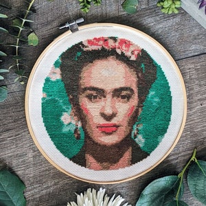 May include: A cross-stitch portrait of Frida Kahlo in a wooden embroidery hoop. The artwork features a detailed depiction of Kahlo with a floral crown, set against a green background. The hoop is surrounded by white flowers and greenery.