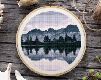 May include: A cross-stitch embroidery hoop with a design of a mountain range reflected in a lake. The mountains are dark blue and the lake is a light blue with a white cloudy sky above.