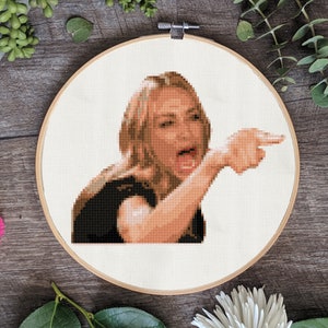 May include: A cross-stitch embroidery of a woman with blonde hair pointing to the right and yelling. The woman is wearing a black shirt and has a surprised expression on her face.