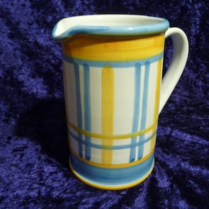 Könnte beinhalten: Handbemalter Keramikkrug mit weißem Griff und Ausguss. Der Krug hat einen weißen Sockel mit gelben und blauen Streifen und Akzenten. Der Rand und der Boden sind blau, und der Krug ist etwa 18 cm hoch.