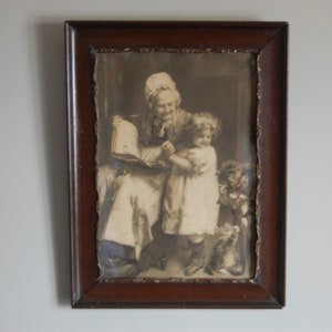 May include: A framed black and white print depicting a seated woman reading to a young child, with a small figure to the right. The print has a torn edge and is set in a dark brown wooden frame.