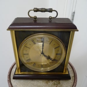 Könnte beinhalten: Eine rechteckige Vintage-Uhr mit goldfarbenem Zifferblatt und römischen Ziffern. Die Uhr hat eine dunkelbraune Holzoberseite und -basis mit goldfarbenen Metallverzierungen. Ein Griff ist oben angebracht. Die Uhr ist etwa 20 cm breit.