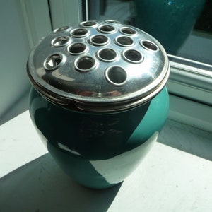 May include: A teal ceramic vase with a silver metal flower frog lid. The lid has multiple circular holes for arranging flowers. The vase is sitting on a white windowsill, with sunlight illuminating the scene. A decorative item for floral arrangements.