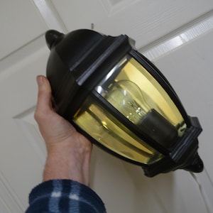 Puede incluir: Luminaria de pared exterior negra con un panel de vidrio transparente y un interior teñido de amarillo. La luz tiene un diseño tradicional con una bombilla visible en el interior. La luminaria está sujeta por una persona.