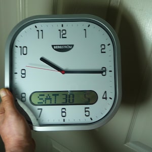May include: A square wall clock with a silver frame and a white face featuring black Arabic numerals. The clock has black hour and minute hands, a thin red second hand, and the brand name Bergstrom printed below the 12. A digital display shows SAT 30.