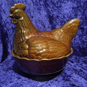 Può includere: Un piatto marrone in ceramica a forma di gallina con coperchio, progettato per assomigliare a una gallina seduta. Il piatto è di colore marrone scuro con finitura lucida. La gallina ha dettagli, tra cui cresta, becco e piume. Il piatto poggia su una base abbinata.