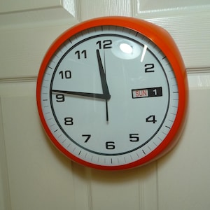 Può includere: Un orologio da parete rotondo con una cornice arancione brillante e un quadrante bianco. L'orologio presenta numeri neri da 1 a 12, lancette nere per ore e minuti, una lancetta dei secondi sottile e una finestrella con la scritta SUN 1.