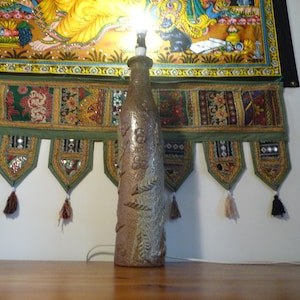 Könnte beinhalten: Braune Keramiklampe in Flaschenform mit geprägten Blatt- und Blumenmustern. Die Zahl "8" ist sichtbar. Die Lampe ist angeschlossen und leuchtet. Im Hintergrund befinden sich farbenfrohe Wandteppiche.