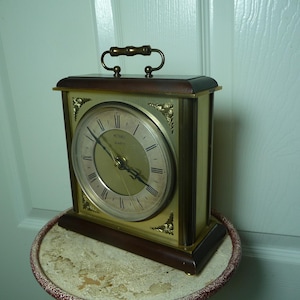 May include: A rectangular Metame Quartz clock with a gold-tone face, Roman numerals, and a dark brown wooden frame. The clock has a handle on top and sits on a round table.