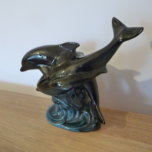Könnte beinhalten: Eine glänzende, dunkelgrüne Keramik-Delfinfigur. Zwei Delfine sind ineinander verschlungen, einer springt aufwärts. Der Sockel zeigt stilisierte Wellen in Türkis. Die Figur steht auf einer Holzoberfläche.